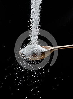 White refined sugar crystals falling down into the wooden spoon at black background