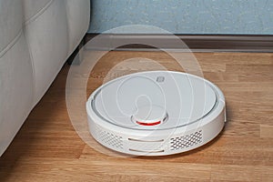 White Robot vacuum cleaner runs in corner near sofa on wood parquet floor. Modern smart cleaning technology housekeeping