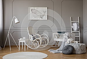 White rocking chair with pillow in the middle of cozy baby room interior with wooden cradle, industrial white lamp and poster in