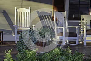 White rocking chairs and potted plants on an open porch in slanting sunlight.