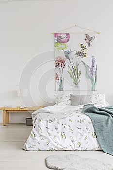 White room interior with double bed, wooden bedside table with lamp and book and material painting hanging on the wa