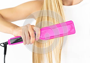 In a white room, a stylish blonde woman with long hair in black straightens her long blonde hair with a pink curling iron
