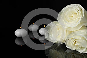 White roses and burning candles on black mirror surface in darkness, closeup with space for text. Funeral symbols
