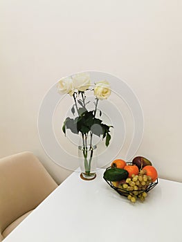 White roses in a glass vase, on a white table with a fruit plate with grapes, avocados, persimmons, mangoes and apples