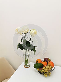 White roses in a glass vase, on a white table with a fruit plate with grapes, avocados, persimmons, mangoes and apples