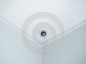 A white round indoor CCTV surveillance camera monitoring security on ceiling in the room corner inside the white clean building.