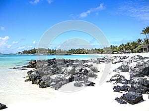 White sandy beach with scattering of rocks in forefront and aqua sea with palm tress in distance