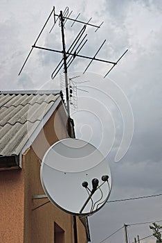 A white satellite dish and an iron television antenna on a brown wall of a house