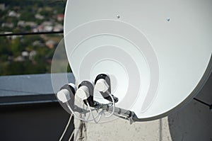 White satellite dish with three converters mounted on residental building rooftop concrete wall. Satellite television