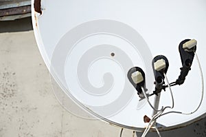 White satellite dish with three converters mounted on residental building rooftop concrete wall. Satellite television