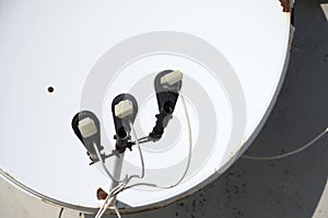 White satellite dish with three converters mounted on residental building rooftop concrete wall. Satellite television
