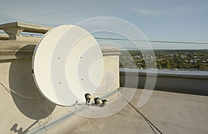 White satellite dish with three converters mounted on residental building rooftop concrete wall. Satellite television