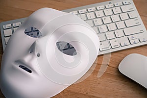 White scary half face mask, keyboard and mouse on wooden table background in dark tone. Hacker, cybercrime, romance scam