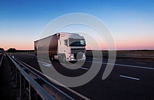 A white semi-trailer truck drives down the highway in the evening with its headlights on. Blue sky and sunset. The mode