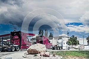 White semi truck pulls two new red semis into fuel service station under stormy skies