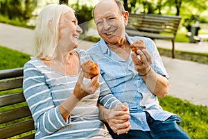 White senior couple smiling and eating muffins while sitting on bench