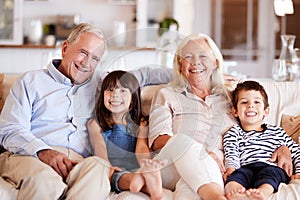 White senior couple and their grandchildren sitting on a sofa together at home smiling to camera