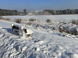 White shades of winter, a snow-covered bench by a frozen pond
