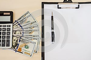 A white sheet of paper on a stand, a pen, dollars and a calculator on the table. View from above.