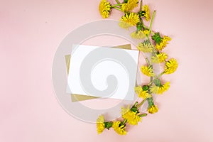 White sheet for text or postcard lies on a pink background, top view. Bright yellow spring dandelions are woven into a wreath and