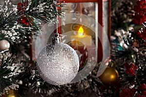 White shiny ball hanging on a branch of a Christmas tree against a red lantern with a candle