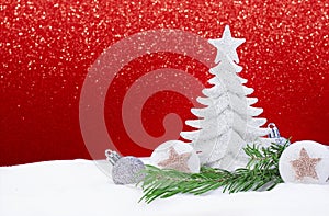 White shiny Christmas tree with silver balls and stars with pine branch on snow on side, behind sparkle red background. New Year