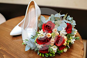 White shoes of the bride with a bouquet of red roses lie on the table in the hotel room