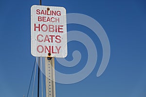 White sign on a metal post that says Sailing Beach Hobie Cats only against a clear cloudless blue sky