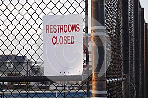 White sign with red lettering that says, Restrooms Closed on a metal chain link fence by a playground