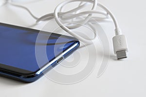 A white signed usb type-c connector lies on a light surface next to a blue smartphone. Connecting and charging gadgets. The