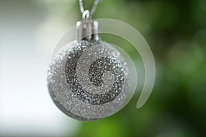 White silver Christmas ball ornament hanging on a blur green tree bokeh light background. Closeup of single Christmas ball