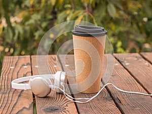 White and silver headphones and a paper Cup of coffee on the wooden table.