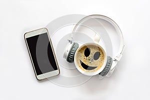 White smartphone with blank mock up screen, headphones, cup of coffee with smile face on white background. Top view. Copy space