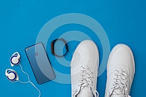 White sneakers, headphones, smart bracelet and blue smartphone on blue background. Sports style. Flat lay.