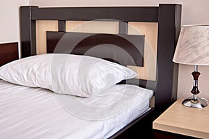 White soft pillow on single bed with brown wooden headboard in hotel bedroom interior, copy space. Classic lamp on bedside table