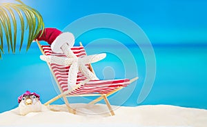 White starfish with Santa\' hat on striped red sun lounger with puffer fish on sandy beach with palm tree behind sea