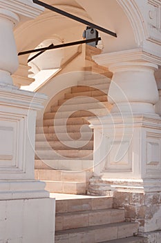 A white stone marble bright classic staircase are leading up with a curve and pillars with an ornament. Sunrays are passing