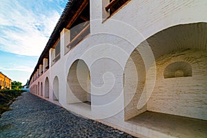 White stone wall of the courtyard with large arches and a wooden roof along the road with masonry in cloudy weather with blue