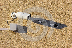 A white street videocamera with cable and a box hangs on a concrete wall and casts a shadow over which the eyes of a spying spy