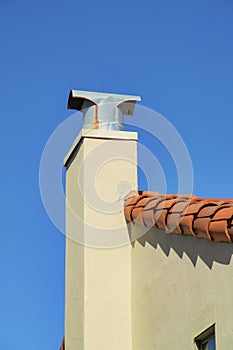 White stucco chimney with metal top with rust marks and red adobe roof tiles on shady roof and house facade in the