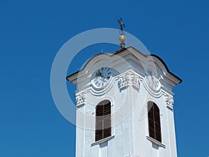 White stucco church clock tower with round clocks and black cross at top.
