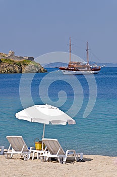 White sunshade with two chairs on a sandy beach with ship anchored by the old roman fortress in a background, Sithonia