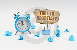 On a white surface, an alarm clock, an office clip, crumpled paper and a cardboard sign with the text -TIME TO NEGOTIATE