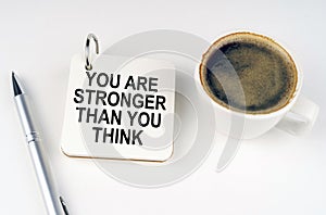 On a white surface, a cup of coffee, a pen and a notepad with the inscription - You Are Stronger Than You Think