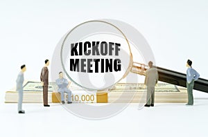 On a white surface, a dollar, figurines of people and a magnifying glass with the inscription -Kickoff Meetin