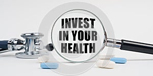 On a white surface lie pills, a stethoscope and a magnifying glass with the inscription - INVEST IN YOUR HEALTH