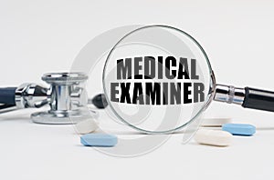 On a white surface lie pills, a stethoscope and a magnifying glass with the inscription - MEDICAL EXAMINER