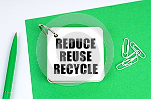 On a white surface lies a green sheet of paper, a pen and a notebook with the inscription - Reduce Reuse Recycle