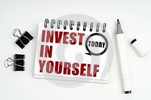 On a white surface lies a marker, clips and a notebook with the inscriptions Today and INVEST IN YOURSELF