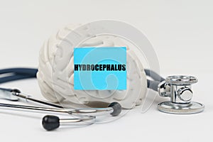 On a white surface next to the stethoscope lies a brain on which a sticker with the inscription - Hydrocephalus